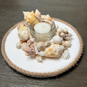 May include: A decorative candle holder featuring a white base with a rope border. The holder is adorned with various seashells in shades of white, beige, and brown, surrounding a small glass jar holding a white candle.