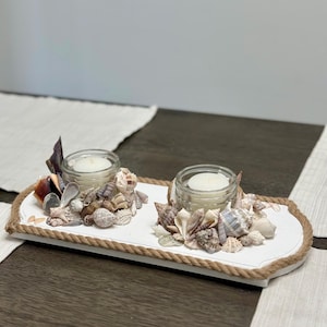 May include: A white, rectangular candle holder with two glass jars holding white candles. The holder is decorated with various seashells and rope trim. The candles are surrounded by an assortment of seashells in shades of white, brown, and tan.
