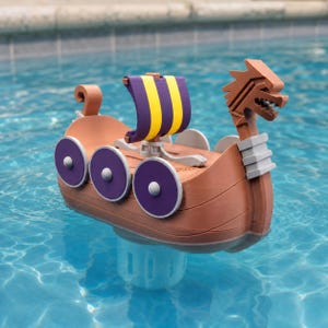 May include: A brown toy Viking ship floating in water, featuring a dragon head prow, a purple and yellow striped sail, and three purple circular shields mounted on each side of the hull. The ship has a light gray accent piece near the dragon head.