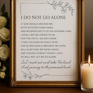 May include: A framed print with the text "I DO NOT GO ALONE" in a gold frame. The print includes a poem with floral accents. A lit candle and flowers are visible in the background.