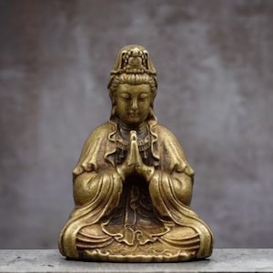 K&ouml;nnte beinhalten: Kleine Messingstatue im antiken Stil einer sitzenden Figur mit gefalteten H&auml;nden. Die Figur tr&auml;gt ein drapiertes Gewand und eine Kopfbedeckung. Die Statue hat ein verwittertes, gealtertes Aussehen, was darauf hindeutet, dass es sich um ein antikes oder Vintage-St&uuml;ck handelt.