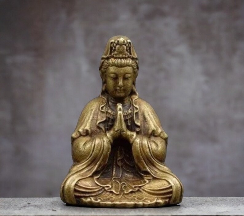 K&ouml;nnte beinhalten: Kleine Messingstatue im antiken Stil einer sitzenden Figur mit gefalteten H&auml;nden. Die Figur tr&auml;gt ein drapiertes Gewand und eine Kopfbedeckung. Die Statue hat ein verwittertes, gealtertes Aussehen, was darauf hindeutet, dass es sich um ein antikes oder Vintage-St&uuml;ck handelt.