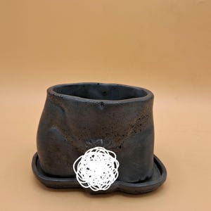 May include: A dark gray ceramic planter with a matching saucer. The planter has a unique, textured surface and an irregular shape. The saucer is attached to the bottom of the planter. The background is a solid tan color.
