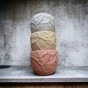May include: Three stacked concrete planters with leaf designs. The top planter is light gray, the middle planter is light yellow, and the bottom planter is light pink.