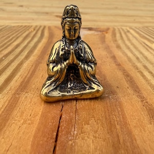 K&ouml;nnte beinhalten: Kleine Messingstatue im antiken Stil, die eine sitzende Figur mit gefalteten H&auml;nden zeigt. Die Figur tr&auml;gt eine detaillierte Kopfbedeckung und flie&szlig;ende Gew&auml;nder. Die Statue ist etwa 5 cm hoch und hat eine verwitterte Oberfl&auml;che.