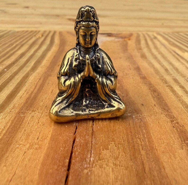 K&ouml;nnte beinhalten: Kleine Messingstatue im antiken Stil, die eine sitzende Figur mit gefalteten H&auml;nden zeigt. Die Figur tr&auml;gt eine detaillierte Kopfbedeckung und flie&szlig;ende Gew&auml;nder. Die Statue ist etwa 5 cm hoch und hat eine verwitterte Oberfl&auml;che.