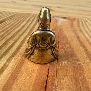 K&ouml;nnte beinhalten: Kleine Buddha-Statue aus Messing im antiken Stil in sitzender Position. Die Figur hat einen glatten, abgerundeten Kopf und ist mit detaillierten Gew&auml;ndern drapiert. Die Statue steht vor einem h&ouml;lzernen Hintergrund.