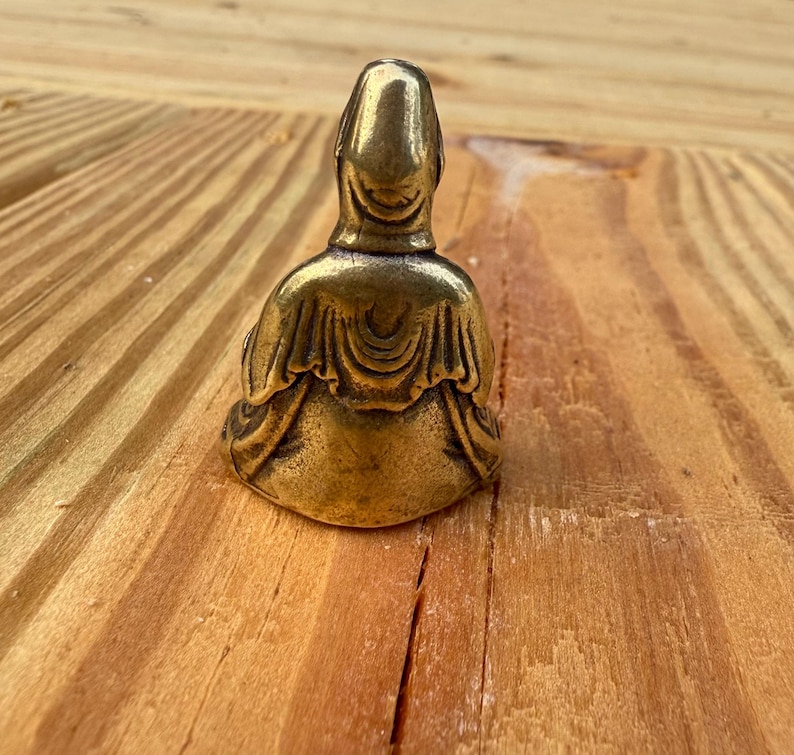 K&ouml;nnte beinhalten: Kleine Buddha-Statue aus Messing im antiken Stil in sitzender Position. Die Figur hat einen glatten, abgerundeten Kopf und ist mit detaillierten Gew&auml;ndern drapiert. Die Statue steht vor einem h&ouml;lzernen Hintergrund.