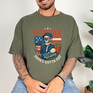 Può includere: T-shirt verde oliva con un motivo a scheletro con occhiali da sole e berretto, che tiene in braccio un bambino e una bevanda. La maglietta mostra il testo "DAD TAX" e "PAPA'S GOTTA EAT" in un design patriottico.