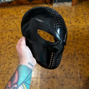 Op de afbeelding: Een zwart plastic masker met een spinnenwebontwerp en een gestructureerd oppervlak. Het masker heeft een grote opening voor de ogen en een kleinere opening voor de mond.