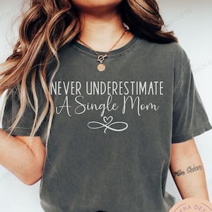 May include: A dark gray t-shirt with the text "Never underestimate a single mom" in white lettering. The shirt has a heart symbol with a swirl design.