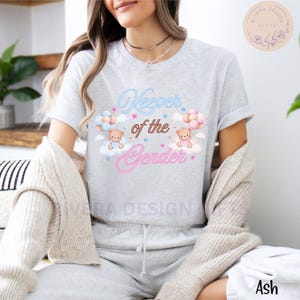 May include: A gray t-shirt with the text "Keeper of the Gender" in pink and blue neon lettering. Two teddy bears are holding blue and pink balloons, floating above clouds with stars.