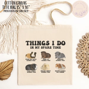 May include: A beige canvas tote bag with the text "THINGS I DO IN MY SPARE TIME" printed in black. The bag features six illustrations of guinea pigs with text describing activities related to guinea pigs, such as "Hang out with guinea pigs", "Look at guinea pigs", "Research guinea pigs", "Talk about guinea pigs", "Dream about guinea pigs", and "Watch guinea pig videos".