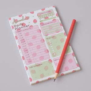 May include: A strawberry-themed daily planner with a red pencil. The planner features a pink and green color scheme with strawberry illustrations. It includes sections for scheduling, mood tracking, priorities, and notes. The text "Amanda's Daily Planner" is visible.