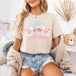 May include: A beige t-shirt with the text "Keeper of the Gender" in pink and blue lettering. Two teddy bears are holding balloons, one blue and one pink, above the text. The shirt is being worn by a person with long brown hair.