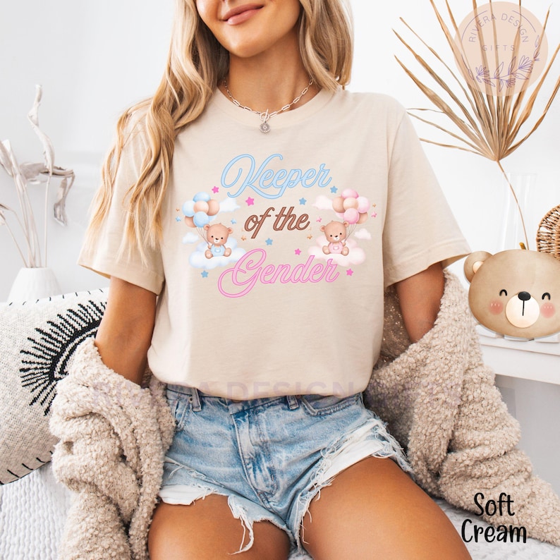 May include: A beige t-shirt with the text "Keeper of the Gender" in pink and blue lettering. Two teddy bears are holding balloons, one blue and one pink, above the text. The shirt is being worn by a person with long brown hair.