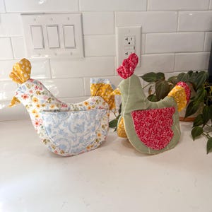 May include: Two fabric chicken-shaped pot holders. One is light blue with a floral pattern, the other is green with a red floral pattern. Both have yellow accents and are sitting on a white countertop.