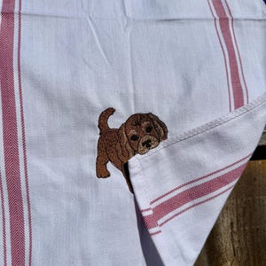 Peut inclure: Un torchon de cuisine blanc avec un motif à chevrons et des rayures verticales rouges. Un chiot marron brodé est situé sur le tissu, semblant regarder derrière un coin replié du torchon.