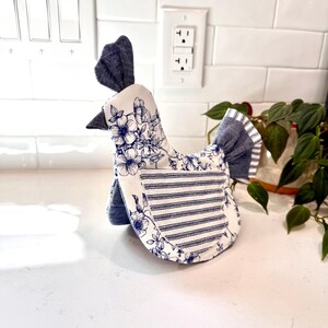 May include: A fabric chicken-shaped kitchen decor item. The chicken has a blue and white floral pattern on the front and a blue and white striped pattern on the sides. The comb and tail are made of solid blue fabric.
