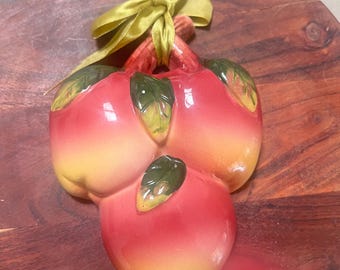 May include: A ceramic apple cluster with red and yellow coloring, green leaves, and a gold ribbon. The decorative piece is set on a dark wooden surface.