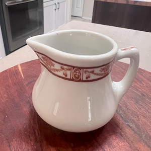 May include: Cream-colored ceramic creamer with a rounded body and a handle. A decorative band of brown floral and ribbon designs encircles the rim. The creamer sits on a dark wooden surface.