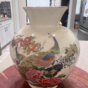 Puede incluir: Un jarrón de cerámica grande, de color crema, con un cuerpo ancho y redondo y un cuello acampanado. El jarrón está decorado con imágenes coloridas de pavos reales, flores y follaje. Las plumas del pavo real son de tonos azules, verdes y marrones.