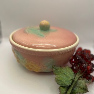 Puede incluir: Un cuenco de cerámica rosa vintage con tapa, con un diseño floral con acentos amarillos y verdes. El cuenco tiene un borde de cuerda alrededor del borde superior.