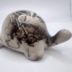 Puede incluir: Figura de tortuga de cerámica con cuerpo beige claro y cabeza y extremidades detalladas. El caparazón de la tortuga presenta un diseño decorativo con rayos negros y blancos y patrones geométricos. La figura mide aproximadamente 15 cm de largo.