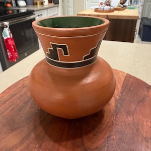 Puede incluir: Un jarrón de cerámica de color terracota con base bulbosa y parte superior acampanada. La parte superior presenta un diseño geométrico en blanco y negro. El interior del jarrón es verde. El jarrón se exhibe sobre una superficie de madera.