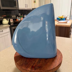 May include: A light blue ceramic vase with a rounded shape, set on a small, round, wooden stand. The vase has a glossy finish and a smooth surface. The wooden stand has a dark brown color and three small metal legs.