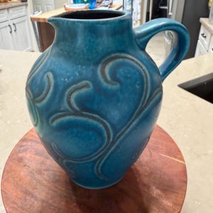 Könnte beinhalten: Türkisblaue Keramikkrug mit dunklem Innenraum und gebogenem Griff. Der Krug hat ein erhabenes, wirbelndes Design in einem dunkleren Türkiston. Er steht auf einem runden Holzständer und zeigt eine rustikale Ästhetik. Der Krug ist etwa 20 cm hoch.