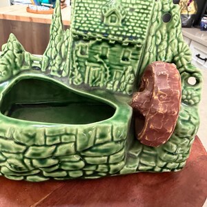 May include: A green ceramic planter shaped like a cottage with a water wheel. The planter has a textured, brick-like base and a dark green interior. The water wheel is a reddish-brown color.