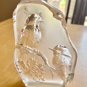 May include: A clear glass sculpture of two birds perched on a branch with leaves. The birds are detailed and realistic, and the sculpture is a beautiful example of glass art.