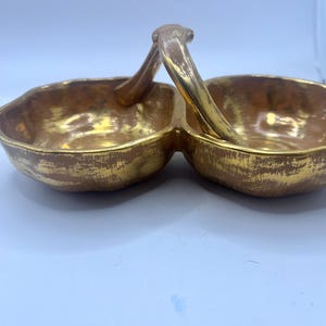 May include: A gold-coloured, two-section serving dish with a handle. The dish has a textured surface and a glossy finish. The handle arches over the two sections, connecting them. The dish is likely for serving snacks or appetizers.