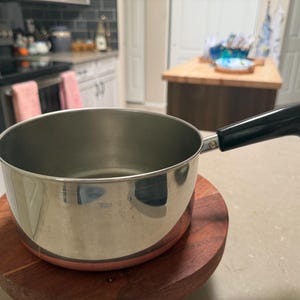 Puede incluir: Una cacerola de acero inoxidable con un asa negra. La olla se encuentra sobre un salvamanteles redondo de madera. El fondo de la cocina incluye una estufa, armarios y una encimera.