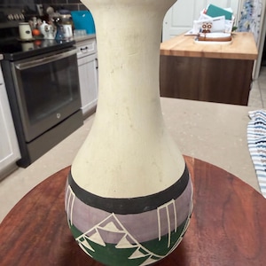 May include: A cream-coloured ceramic vase with a wide base and flared rim. The base has a geometric design with green, purple, black, and white shapes. The vase is displayed on a dark wooden surface.