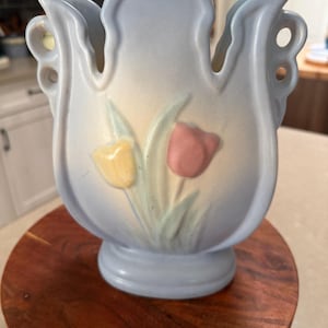 Könnte beinhalten: Hellblaue Keramikvase mit Tulpenmuster. Die Vase hat einen ausgestellten oberen Rand mit dekorativen Ausschnitten und einen Sockel. Die Tulpen sind in Gelb und Rosa mit grünen Stielen und Blättern bemalt. Die Vase steht auf einer runden Holzoberfläche.