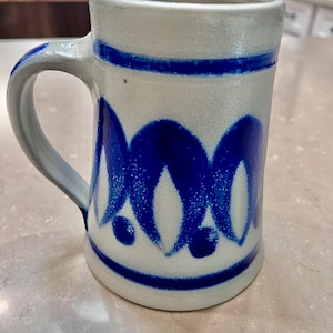 Puede incluir: Una jarra de cerveza de cerámica blanca con un diseño azul y blanco. La jarra tiene un asa y una boca ancha.