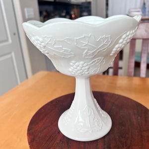 Puede incluir: Un tazón de pedestal de vidrio de leche blanco con un diseño de vid. El tazón tiene un borde festoneado y una base con pie.