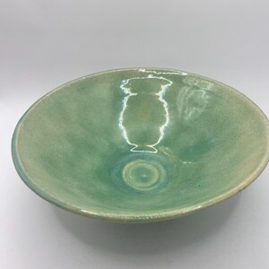 May include: A handmade ceramic bowl with a glossy, seafoam green glaze. The bowl has a wide, shallow design with a slightly raised rim. The interior features a subtle spiral pattern, adding visual interest to this functional art piece.