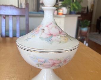 May include: A white ceramic candy dish with a floral design. The dish has a lid and a pedestal base. The flowers are pink, yellow, and blue.