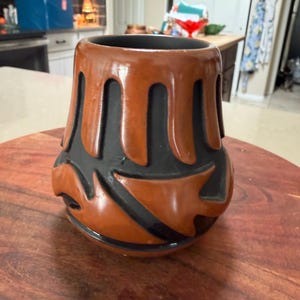 May include: A brown and black ceramic vase with a unique, carved design. The vase has a wide base and a narrower top, with a series of vertical, curved cutouts. The color scheme is a warm brown with black accents, creating a striking contrast.
