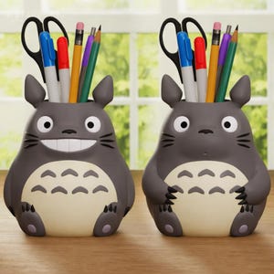Puede incluir: Dos organizadores de escritorio grises con forma de Totoro, cada uno con bolígrafos, lápices y tijeras. Los organizadores tienen una panza beige claro y una cara sonriente. El fondo muestra una ventana.