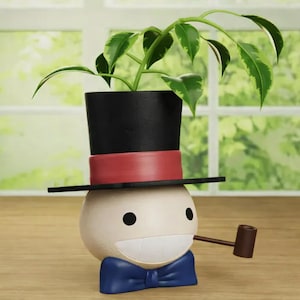 Pode incluir: Um vaso de plantas extravagante em forma de personagem de desenho animado usando uma cartola preta com uma faixa vermelha, uma gravata borboleta azul e um cachimbo. O vaso contém uma planta verde com folhas variegadas. O personagem tem um rosto sorridente.