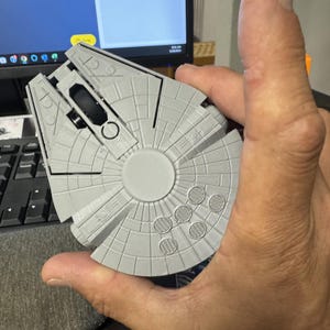 May include: A grey computer mouse in the shape of the Millennium Falcon spaceship. The mouse features a circular design with detailed paneling and a black scroll wheel. The mouse is held in a person's hand.