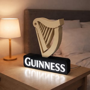 Peut inclure: Lampe en forme de harpe crème avec le mot "Guinness" en lettres blanches sur une base noire. La lampe est posée sur une table de chevet en bois à côté d'une lampe de table allumée.