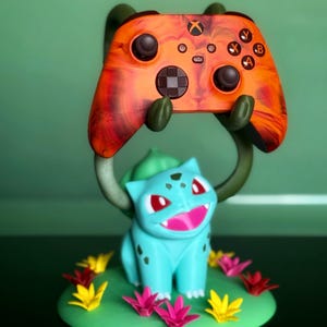May include: An Xbox controller in orange and black, held by a Bulbasaur stand. The controller has black buttons and an Xbox logo. The Bulbasaur figure is light blue with red eyes and a pink mouth, surrounded by colourful paper flowers.