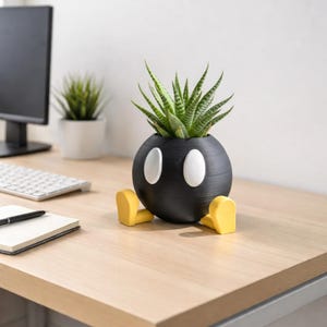 Könnte beinhalten: Ein schwarzer, kugelförmiger Pflanztopf mit weißen ovalen Akzenten und gelben Füßen, der einer Cartoon-Bombe ähnelt. Er enthält eine grüne Sukkulentenpflanze. Der Pflanztopf steht auf einem hellen Holzschreibtisch.