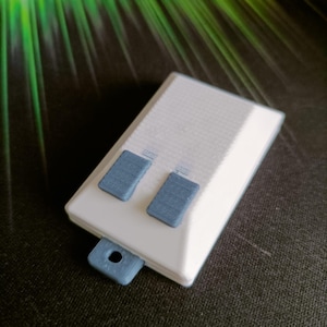 Può includere: Un oggetto rettangolare bianco stampato in 3D con due pulsanti rettangolari blu e una linguetta blu con un foro. La superficie superiore presenta un motivo strutturato. Probabilmente si tratta di un piccolo dispositivo o componente elettronico.