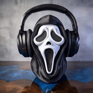 May include: A black and white Scream mask with a black hood, holding a pair of black headphones. The mask has a white face with black eye sockets and a wide open mouth. The headphones are over the mask's head.